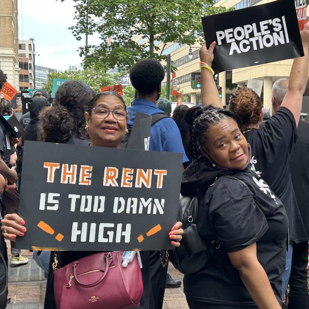 two one northside leaders at a protest with a sign that says the rent is too damn high