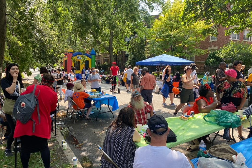 Wide shot of a northside block party. There are tables with people sitting behind them, lots of adults and kids walking around, a bounce house in the distance, and a canopy of trees above