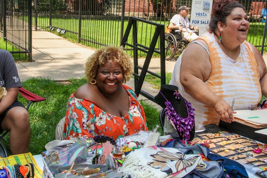 A table is covered with crafts and jewelry. Behind the table are 4 people, 3 of them close to the table, and just 2 of those three can we see their faces. One person facing the camera is wearing an orange blouse and is smiling big. The person next to her is talking to someone else who is off camera.