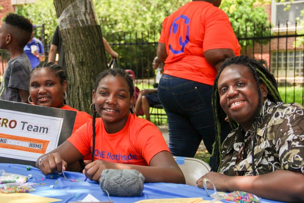 Three smiling people making friendship bracelets and holding up a sign saying "SRO Team, Bring Chicago Home." Two of these people are wearing orange "ONE Northside" t-shirts, as is a person standing in the background. 