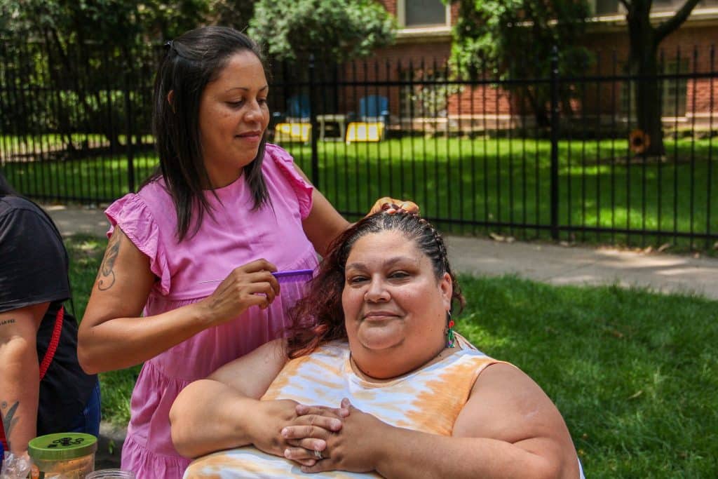 Two people sit in the foreground. One is standing and has a comb in her hand. The other is sitting, and having her hair braided. grass and a sidewalk are in the background. 