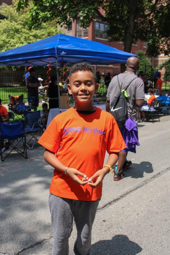 Boy is standing and smiling at the camera with a "ONE Northside" orange shirt. People, tables and a pop-up tent are in the background.