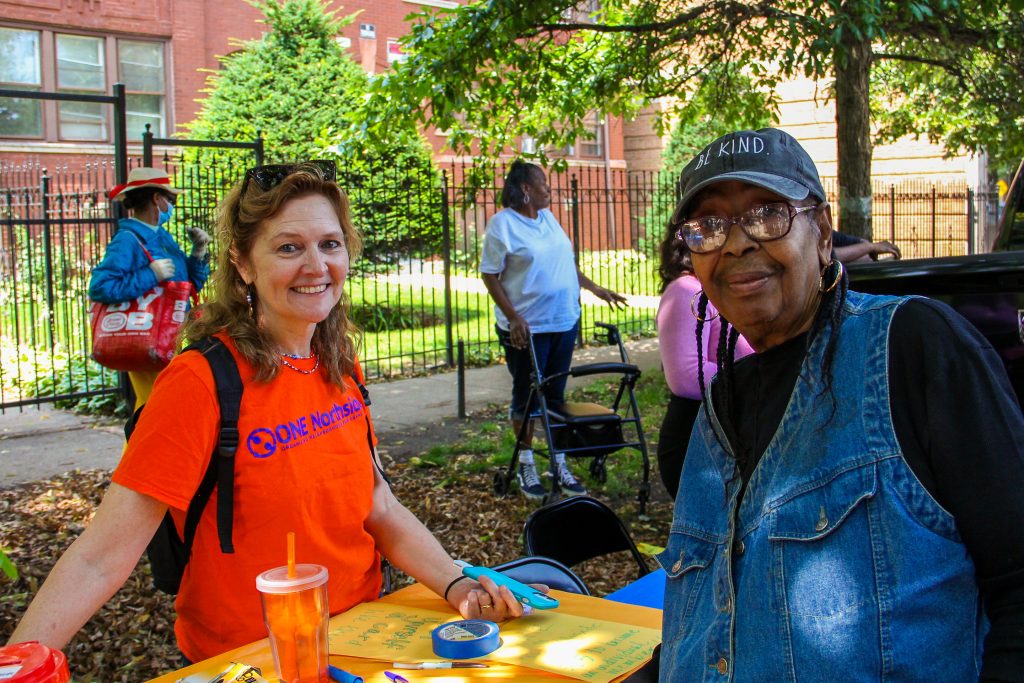 Two people smile into the camera. One is wearing an orange "ONE Northside" t-shirt. There is a table between them, with raffle tickets and a list of giveaway items. 