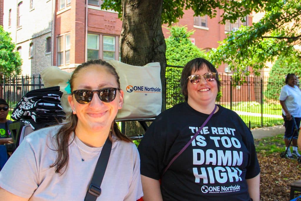 Two smiling faces, both wearing sunglasses, and one is wearing a "The Rent is Too Damn High" t-shirt. Behind them, a rack with t-shirts and a tote bag saying "ONE Northside"