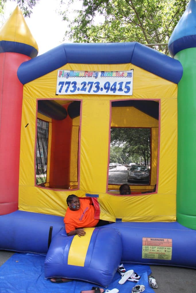 Large red, yellow, blue and green bouncy house , with two kids. One kid is looking through the window and smiling, the other is halfway in and halfway out of the bouncy house door, also smiling. Shoes lay on the ground near the entrance.