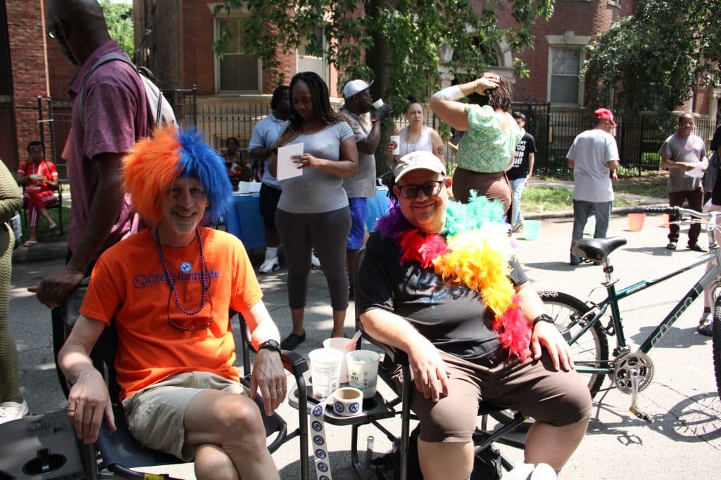 Two men sit in folding chairs, with other folks milling behind them. One man has an orange and blue wig, the colors of ONE Northside, and is wearing an orange ONE Northside t-shirt. The other man is wearing a rainbow feater boa. 