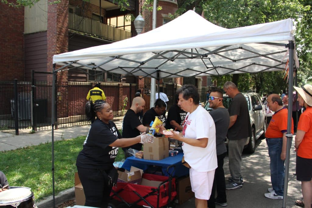 White pop-up tent, with a table underneath for hot dogs and fixins. Folks with "The Rent is too damn high" stand on one side dishing out food, neighbors receive food on the other side.