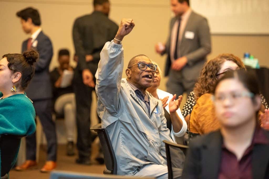 In the center of the shot is a man triumphantly holding up an arm in celebration, and smiling. He is wearing grey pants and a grey jacket and glasses. He is surrounded by people, who are mostly out of focus.