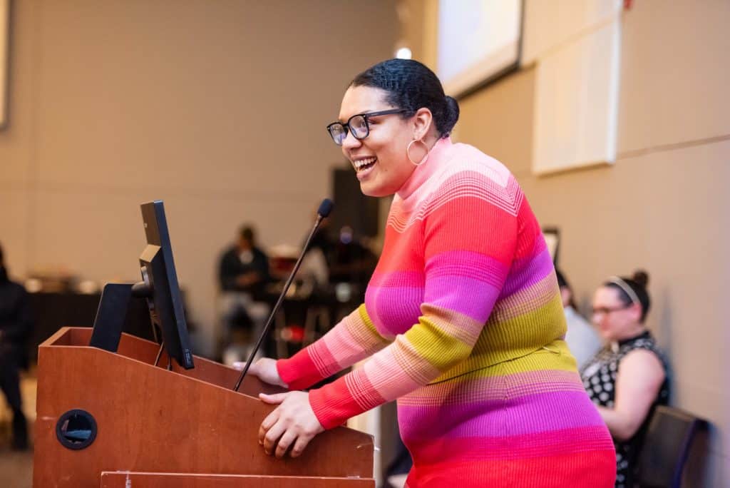 Alderwoman Amanda Clay stands at the podium wearing a long sleeve dress that is red, pink, green, and purple, hoop earrings, a bun and glasses. She is smiling. Behind her we can see Rev Lindsey Joyce in a black and white dress.