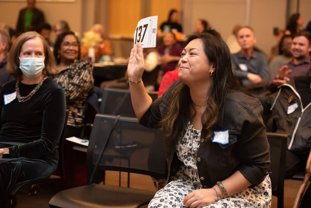 A woman smiles as she holds up a card with a number on it, as people smile in the background.