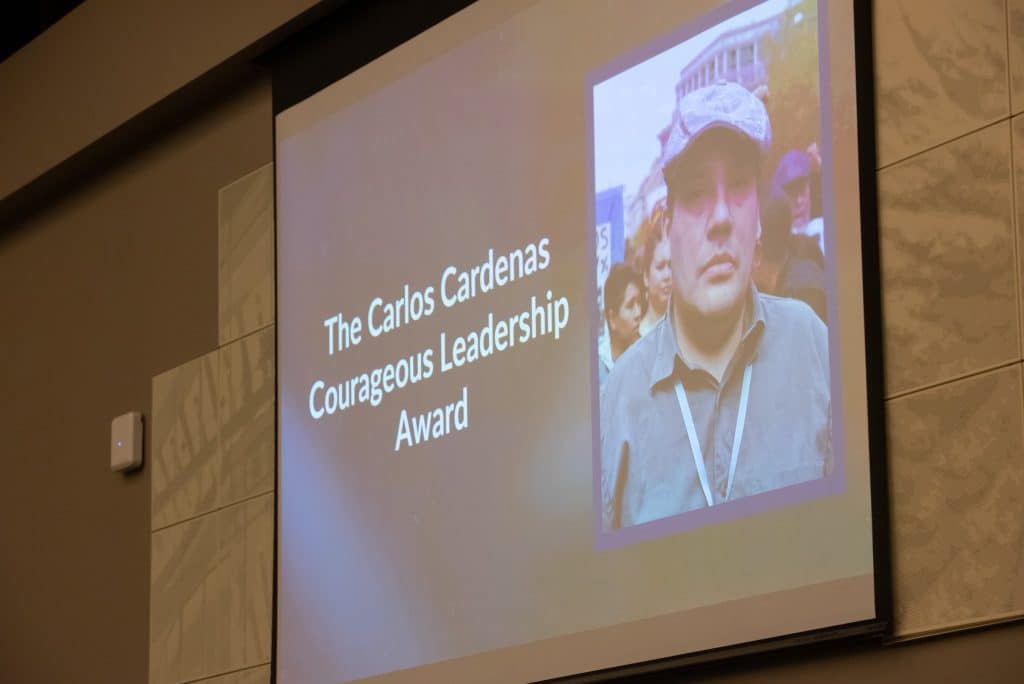 A screen with the words "The Carlos Cardenas Courageous Leadership Award" and an image of a man looking into the camera wearing a hat, button-up and lanyard, and a crowd of people behind him.