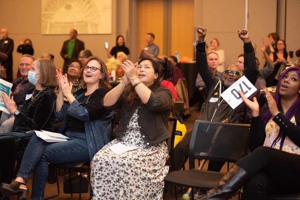 Rows of people in chairs smile, clap and raise their arms in celebration. People can be seen standing at the far back wall, who are also clapping.