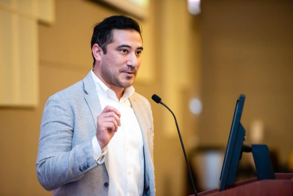Jesse Hoyt, a man in a button up and blazer stands at the podium and talks into the microphone. He is smiling and gesturing.