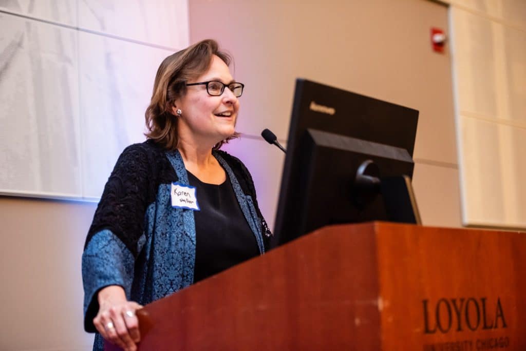 A woman stands at the podium, she is wearing a black and blue dressy outfit, black glasses, and her shoulder-length hair is down. She is smiling as she talks.