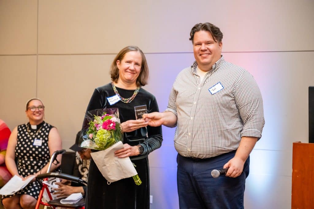 A tall man in a button up shirt holds a glass award with a woman in a black dress holding flowers.