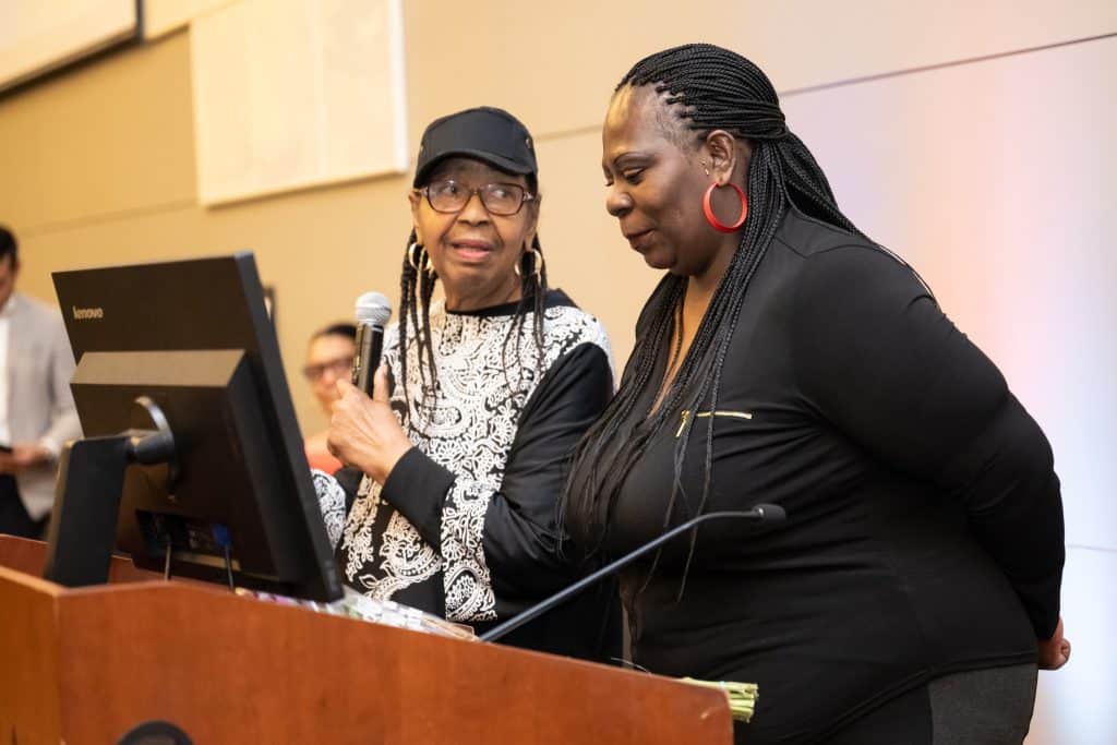 Two women stand at the podium. One is smiling and talking while looking at the other woman, who is smiling and looking down.