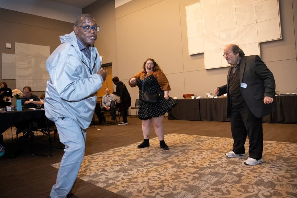 3 people dance, while other folks sit in the background chatting. Of the dancers, two are smiling and looking into the camera.