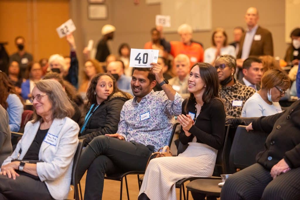 A crowd of people during the paddle raise. Some are sitting in the front, others are behind them in the back. Most people are smiling, and a few people are holding up cards with numbers.