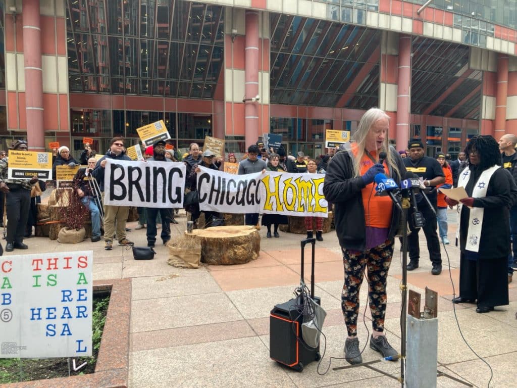 Reina speaks into a microphone in front of the Thompson Center with a big crowd behind her holding signs and banners