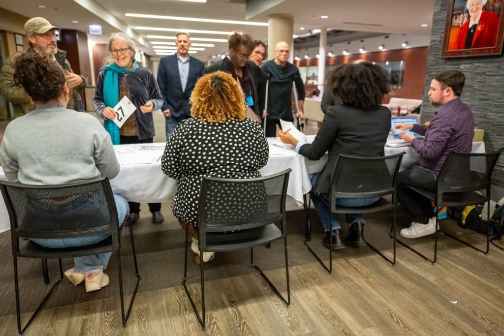 A long check-in table with a table cloth. On one side of the table are four chairs, each with a person sitting, with their back to the camera. On the other side, people are standing and checking in or waiting to check in.
