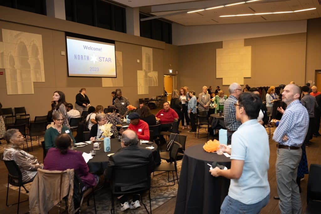 A large ballroom with standing tables, tables and chairs for sitting, and chairs in rows. People are standing and sitting at tables, and in the chair rows. People are dressed nicely and most are wearing name tags. On the wall, a screen reads "Welcome! North Star 2023"