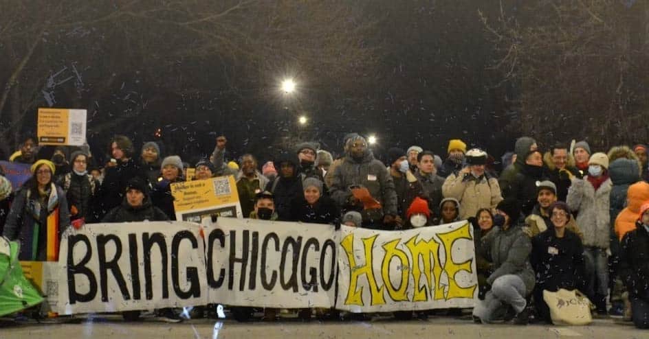 Banner of a few dozen people holding a sign with black and yellow letters that says Bring Chicago Home