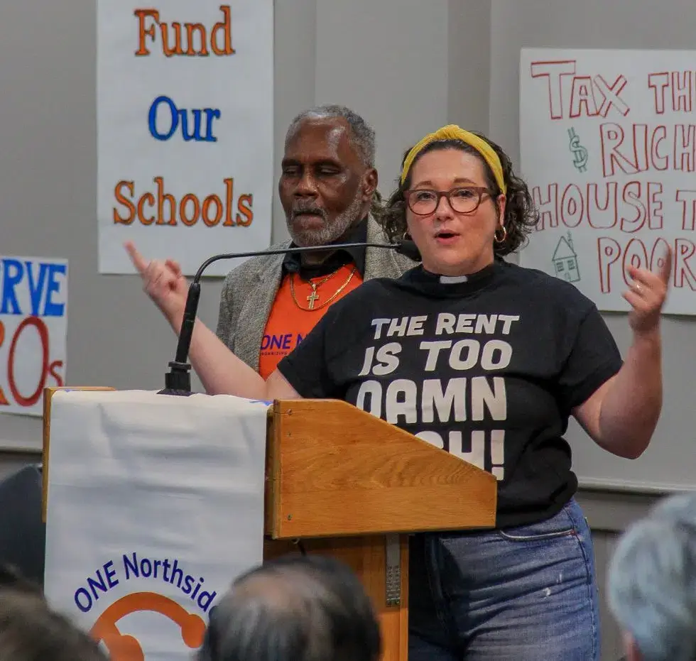 Lindsey at the podium with Anthony just behind her, protest signs on the wall behind them.