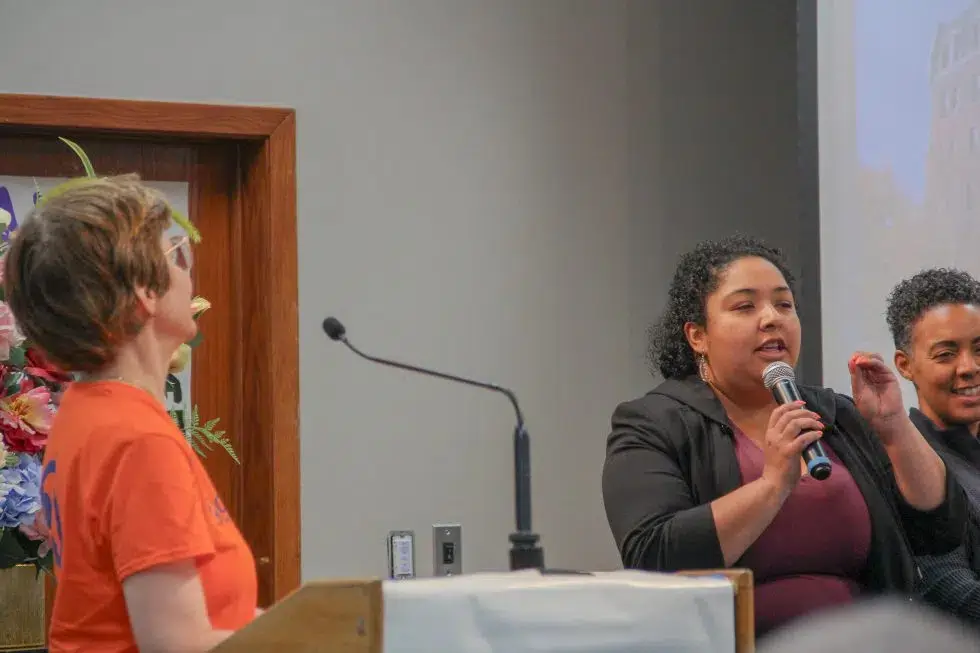 Sue at the podium turns to watch Commissioner Castaneda speaking into a microphone addressing the audience