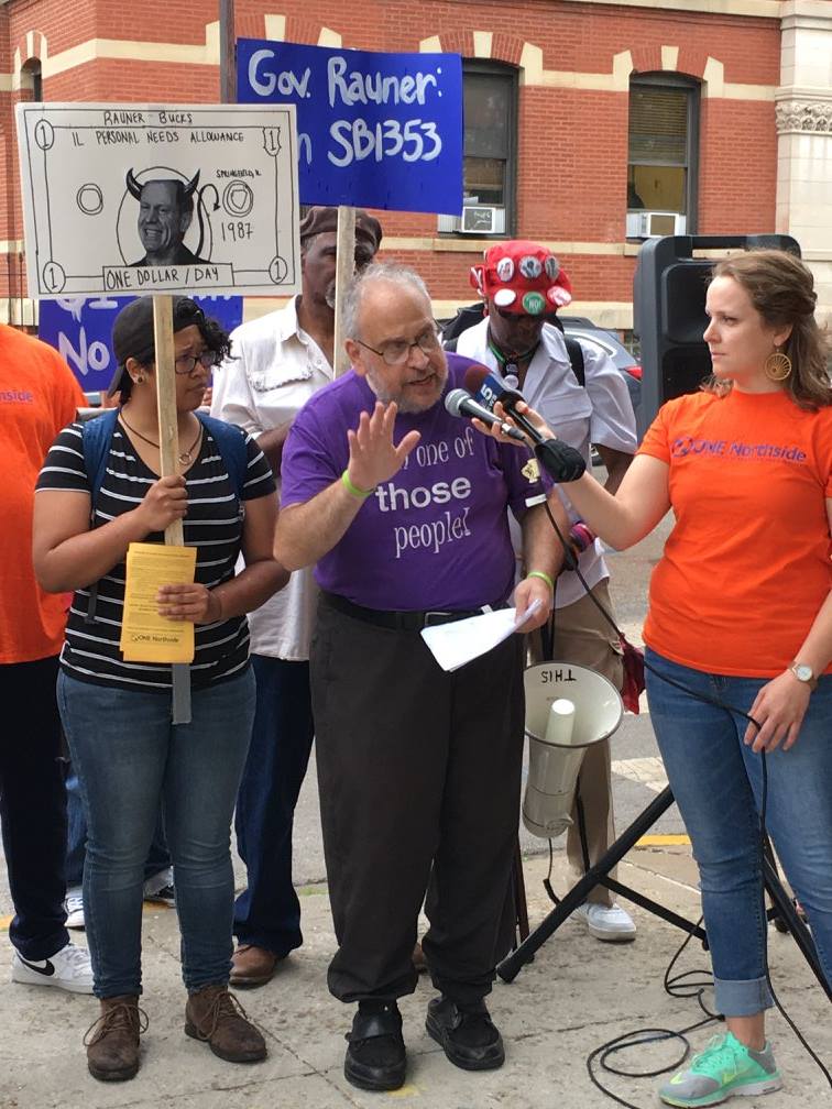 a speaker at a microphone with supporters in the background with signs