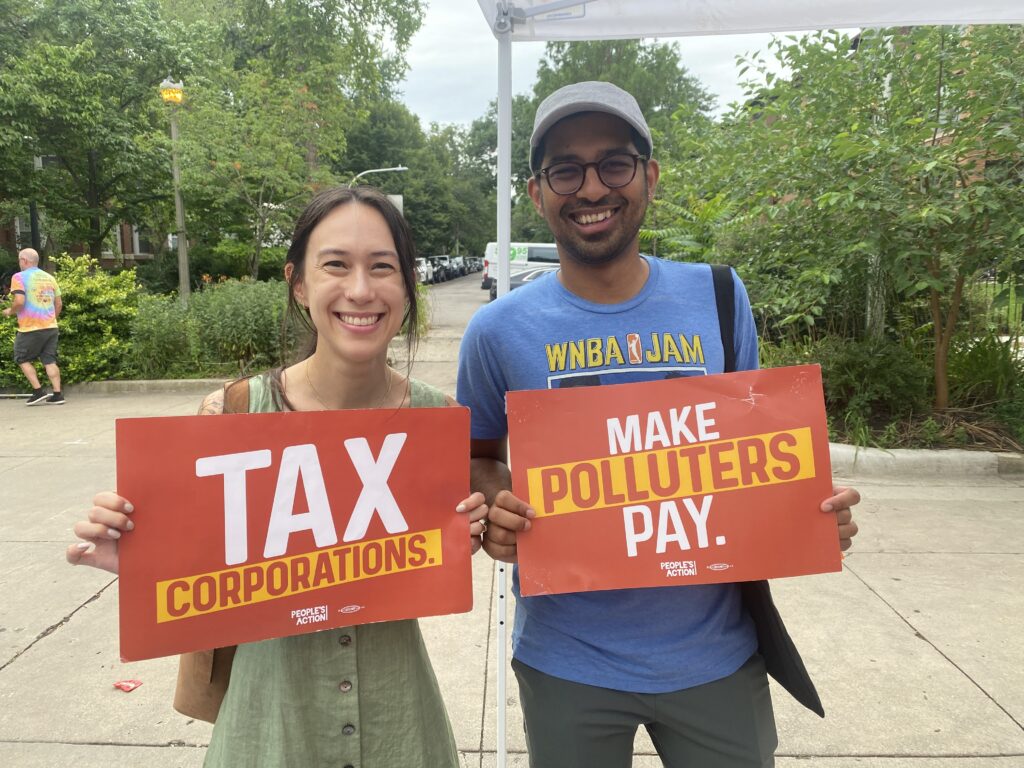 A woman and man holding up two different signs one saying "tax the rich" the other saying "make polluters pay" both smiling