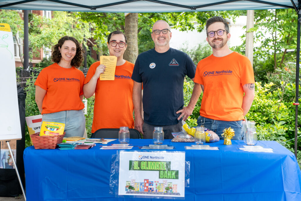 There are 3 men and a younger woman standing behind the IL Climate Bank table at our Back to School Summer Bash event with one man holding up some flyers