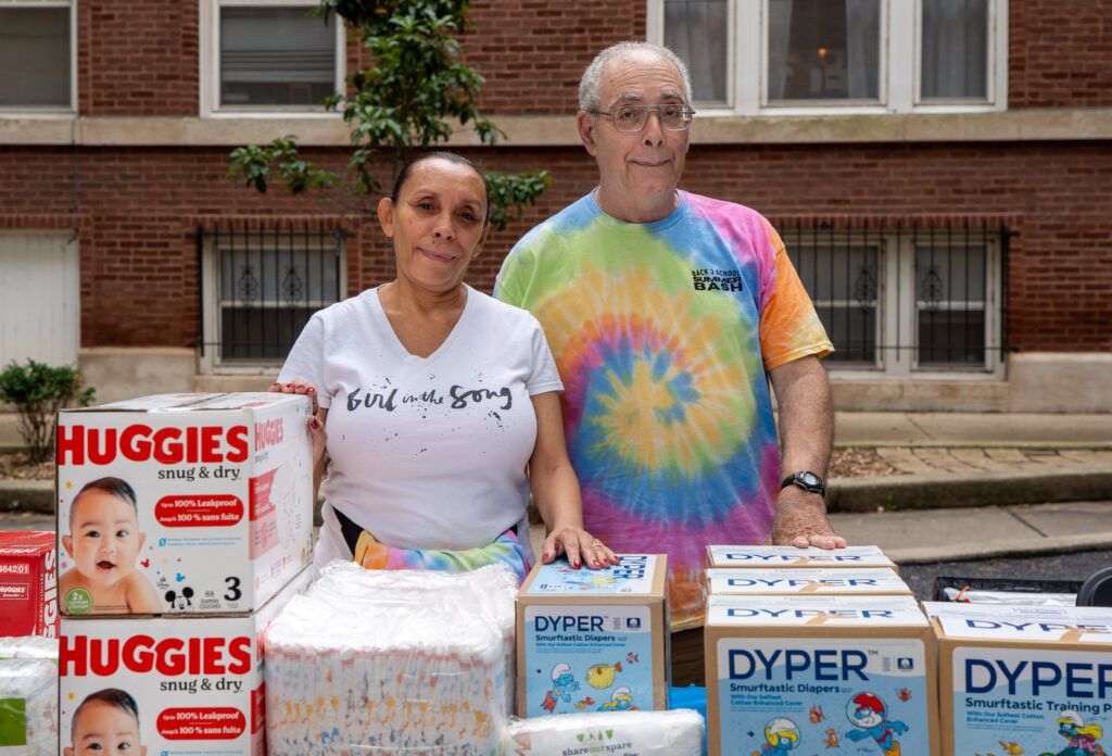 An older woman and an older man in a tye dye t-shirt standing behind a table of diapers