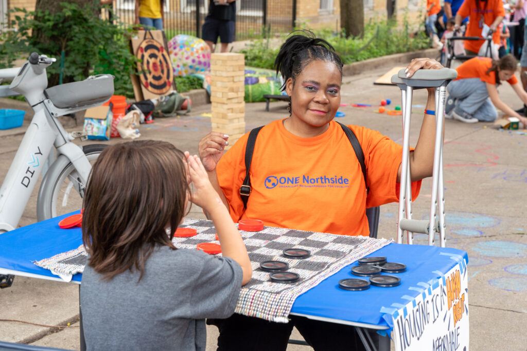 A woman in a ONE Northside t-shirt smiling while playing chess with a younger boy