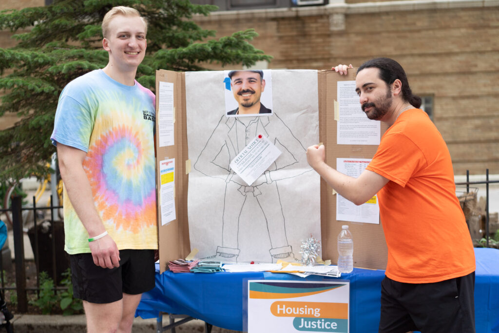 A board with a man face cut out and an eviction notice pined to his body. Two men around this poster with one in an orange t-shirt holding up a fist
